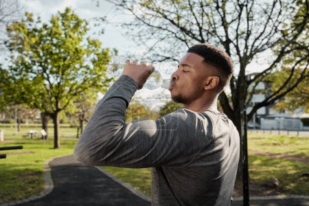 Foto de Joven hombre negro usando ropa deportiva con los ojos cerrados bebiendo agua de la botella en el parque - Imagen libre de derechos