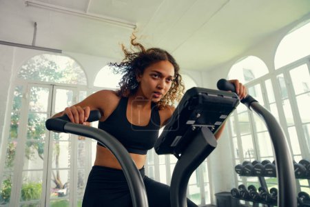 Photo for Focused young multiracial woman wearing sports clothing cycling on exercise bike at the gym - Royalty Free Image