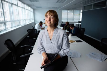 Téléchargez les photos : Joyeux jeune femme d'affaires portant des vêtements d'affaires assis sur le bureau avec tablette numérique dans le bureau de l'entreprise - en image libre de droit