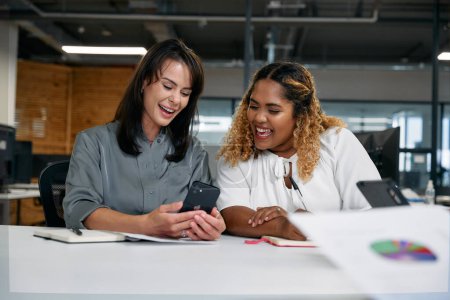 Téléchargez les photos : Deux femmes d'affaires multiraciales heureuses souriant tout en utilisant un téléphone intelligent lors d'une réunion dans un bureau d'entreprise - en image libre de droit