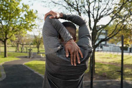Photo for Rear view of young black man wearing sports clothing doing stretches in park - Royalty Free Image