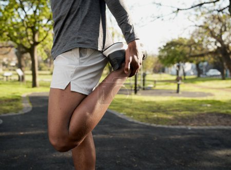 Foto de Primer plano del joven negro que usa ropa deportiva con la pierna levantada mientras hace estiramientos en el parque - Imagen libre de derechos