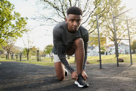 Foto de Joven hombre negro con ropa deportiva mirando a la cámara mientras se arrodilla y ata cordones en el parque - Imagen libre de derechos