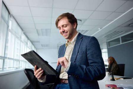 Foto de Feliz joven empresario usando ropa de negocios usando tableta digital por escritorio con compañeros de trabajo en la oficina corporativa - Imagen libre de derechos