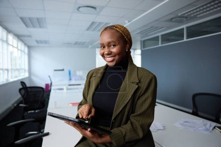 Téléchargez les photos : Confiant jeune femme d'affaires noire portant des vêtements d'affaires en utilisant une tablette numérique par bureau dans le bureau de l'entreprise - en image libre de droit