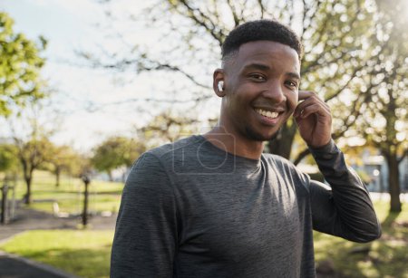 Foto de Joven hombre negro usando ropa deportiva escuchando música con auriculares inalámbricos y sonriendo en el parque - Imagen libre de derechos