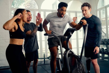 Foto de Adultos jóvenes multiraciales que usan ropa deportiva que motiva a su amigo a montar en bicicleta estática en el gimnasio - Imagen libre de derechos