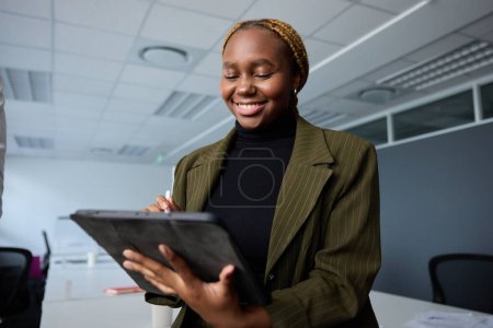 Foto de Joven empresaria con ropa de negocios sonriendo mientras usa tableta digital y pluma digitalizada en la oficina corporativa - Imagen libre de derechos