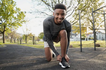 Téléchargez les photos : Heureux jeune homme noir portant des vêtements de sport regardant la caméra tout en s'agenouillant et attachant lacet dans le parc - en image libre de droit