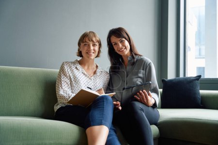 Two happy business women sitting on sofa using digital tablet during meeting in corporate office