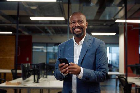 Foto de Feliz hombre negro maduro en ropa de negocios mirando a la cámara mientras se utiliza el teléfono móvil en la oficina corporativa - Imagen libre de derechos