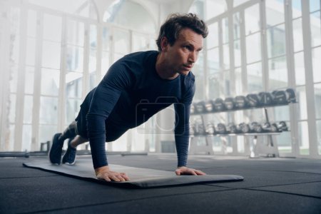 Photo for Young caucasian man wearing sportswear in plank position while doing push-ups on exercise mat at gym - Royalty Free Image