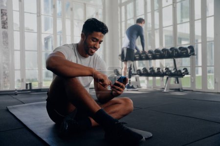 Téléchargez les photos : Deux jeunes hommes multiraciaux portant des vêtements de sport souriants et utilisant un téléphone portable à côté des poids à la salle de gym - en image libre de droit