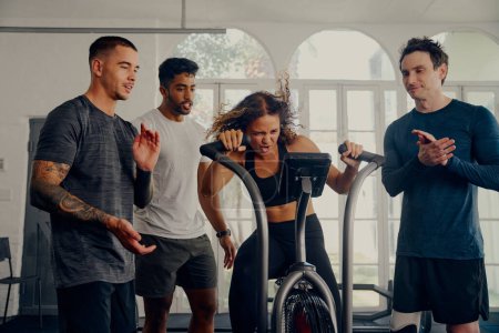 Photo for Multiracial young adults wearing sportswear applauding for friend cycling on exercise bike at the gym - Royalty Free Image