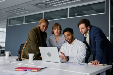 Foto de Cuatro adultos jóvenes que usan ropa de negocios hablando y haciendo gestos con un portátil en el escritorio en la oficina corporativa - Imagen libre de derechos