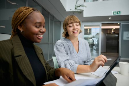 Téléchargez les photos : Deux jeunes femmes d'affaires multiraciales heureuses portant des vêtements d'affaires avec tablette numérique et documents au bureau - en image libre de droit