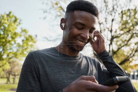 Foto de Joven hombre negro con ropa deportiva escuchando música con auriculares inalámbricos y teléfono móvil en el parque - Imagen libre de derechos