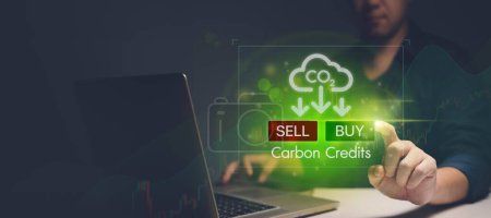In the image, the investor is using a laptop to access the carbon credits market on an online platform. They are raising their hand to use their finger to point at a hologram floating in the air, which is a buy and sell button on the laptop screen, w