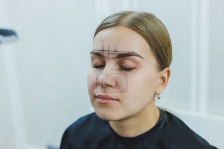 A woman permanent makeup artist draws a sketch of the eyebrows on the face of her client.