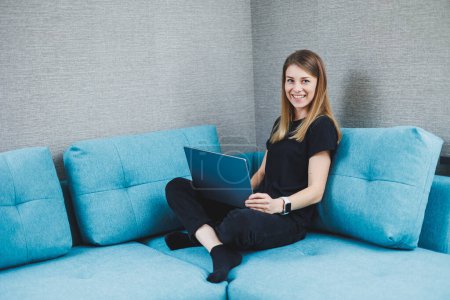 Téléchargez les photos : Une jeune femme est assise sur un canapé et travaille sur un ordinateur portable. Travail à distance. Travail à domicile en ligne - en image libre de droit