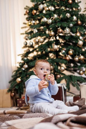 A 2-year-old little boy sits near a Christmas tree decorated with toys. New Year's atmosphere