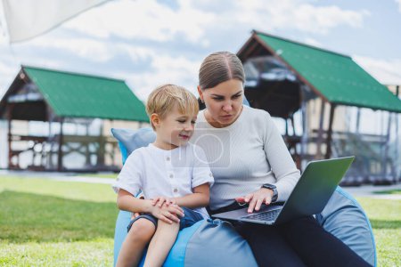 Photo for A mother with a laptop, her son sitting next to her outdoors on a green grass lawn. Rest and work with a child - Royalty Free Image