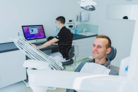 Photo for A young man is sitting in a dental chair, and a dentist is working at a computer. Dentist's appointment - Royalty Free Image