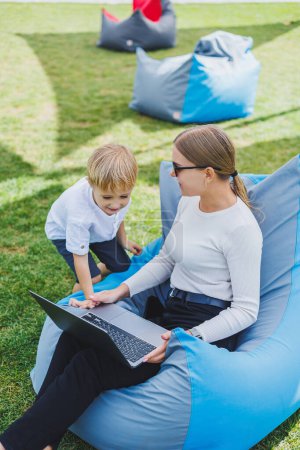 Photo for A mother with her son on a green lawn, a mother on maternity leave is working on a laptop in the park, her son is playing next to her. - Royalty Free Image
