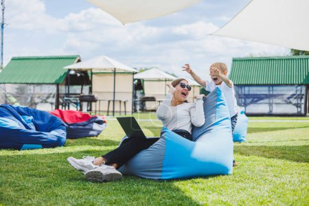Photo for Mother with laptop, her son playing outdoors on green lawn. Rest and work with a child in the park - Royalty Free Image