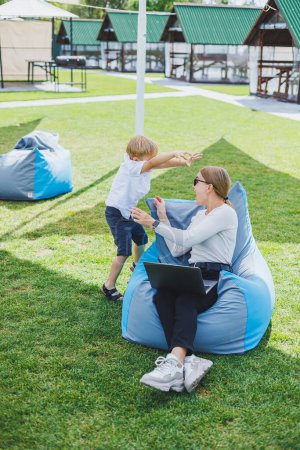 Photo for Mother with laptop, her son playing outdoors on green lawn. Rest and work with a child in the park - Royalty Free Image