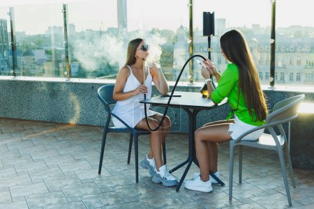 Copines assis sur la terrasse ouverte et la fumée narguilé, se reposer avec des copines
