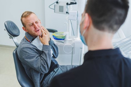 Foto de Un médico dentista joven le dice a un paciente masculino acerca de la atención dental. Un hombre estaba sentado en una silla dental en una cita con el médico. Tratamiento dental moderno - Imagen libre de derechos