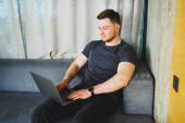 Smiling man in t-shirt sitting on sofa, typing on netbook, working remotely on startup as freelancer, looking at laptop and smiling Poster #647036418