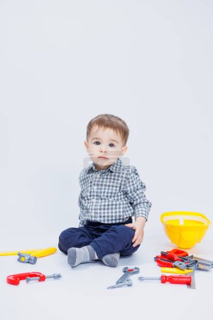 A little boy in a shirt with a helmet and a toy tool. White background. Toy construction tool for children