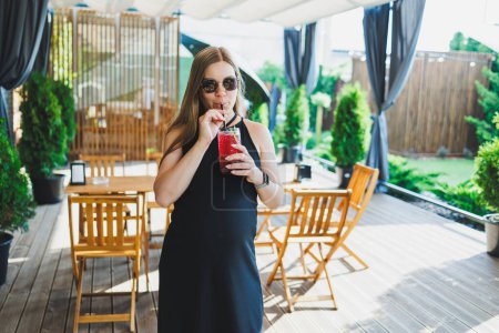 Téléchargez les photos : Une jolie femme enceinte en robe noire sur une terrasse d'été dans un café boit de la limonade fraîche. Une femme enceinte boit de la limonade aux fruits sans alcool - en image libre de droit