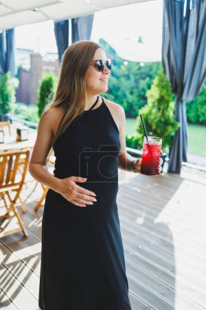 Téléchargez les photos : Une jolie femme enceinte en robe noire sur une terrasse d'été dans un café boit de la limonade fraîche. Une femme enceinte boit de la limonade aux fruits sans alcool - en image libre de droit