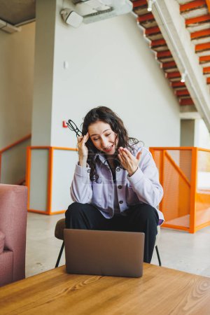 Foto de Estudiante rizada trabajando en la cafetería brillante en línea mirando el ordenador portátil. Trabajo remoto con un portátil en una acogedora cafetería de trabajo - Imagen libre de derechos