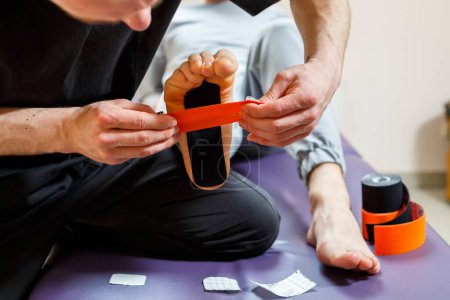 Photo for Male physiotherapist doctor massages the feet of a relaxed man sitting on a stretcher. He uses kinesio tape. Taping the foot - Royalty Free Image