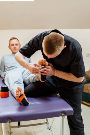 Photo for Male physiotherapist doctor massages the feet of a relaxed man sitting on a stretcher. He uses kinesio tape. Taping the foot - Royalty Free Image