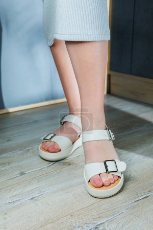 Téléchargez les photos : Sandales en cuir blanc sur les pieds des femmes. Pieds féminins dans des sandales décontractées confortables - en image libre de droit