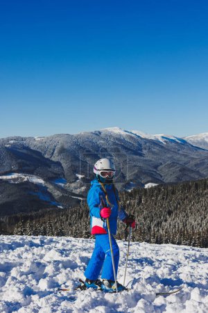 A little girl of 8 years old in a ski suit and on skis stands against the background of snowy mountains. Ski resort