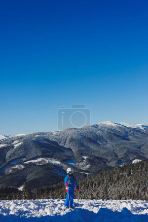 Ein kleines Mädchen von 8 Jahren steht im Skianzug und auf Skiern vor dem Hintergrund verschneiter Berge. Skigebiet
