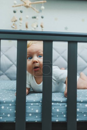 The baby looks through the sides of the crib into the room. A small newborn boy lies in a crib for children. A place for children to sleep
