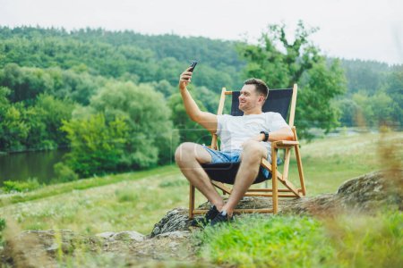 Ein bärtiger Mann sitzt auf einem klappbaren Holzstuhl in der Natur und telefoniert. Allein das Konzept der Erholung in der Natur.