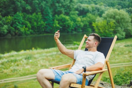 A bearded man sits on a folding wooden chair in nature and talks on the phone. The concept of recreation in nature alone.