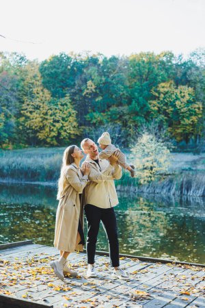 Téléchargez les photos : Un jeune couple heureux avec un petit fils dans les bras dans le parc près du lac. Vacances en famille dans la nature avec un petit fils. Le concept de vacances en famille. - en image libre de droit
