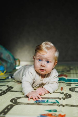 The child crawls on his stomach. A small 6-month-old baby is crawling on the rug and smiling. A boy plays with a toy on the floor.
