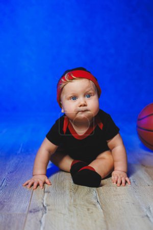 Téléchargez les photos : Portrait d'un bébé de 7 mois en body noir avec un ballon de basket. Du sport pour les enfants. Vêtements en coton pour bébés - en image libre de droit