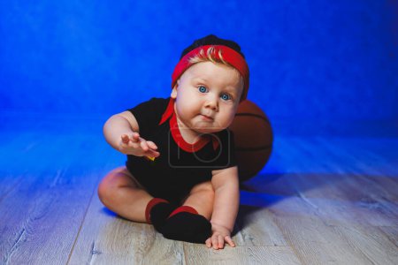 Retrato de un bebé de 7 meses en un traje negro con una pelota de baloncesto. Deportes para niños. Ropa de algodón para bebés