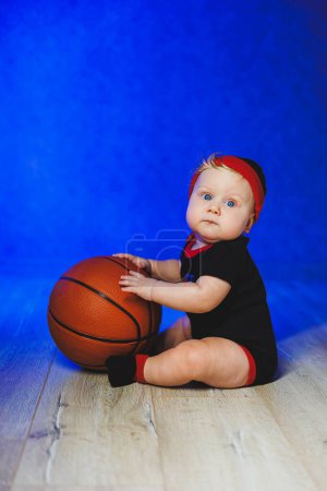 Retrato de un bebé de 7 meses en un traje negro con una pelota de baloncesto. Deportes para niños. Ropa de algodón para bebés